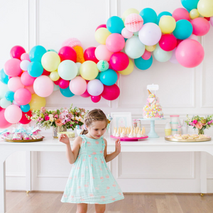 Little girl dancing in front of a white table decorated with ice cream treats. There is a balloon garland behind her with brightly colored balloons.