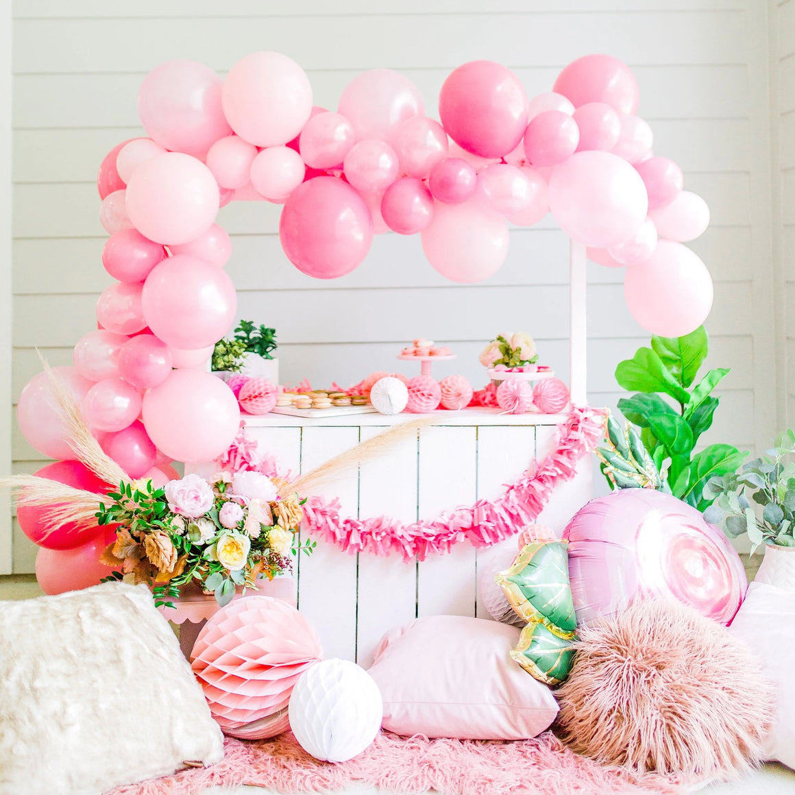 A pretty white lemonade style stand has a beautiful balloon garland draped along the top and down one of the sides. The balloon garland is the perfect mix of pink, pearl pink, and rose colored balloons.