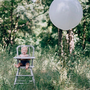 A baby boy sits in a white high chair, next to him is a jumbo 36 inch white balloon.