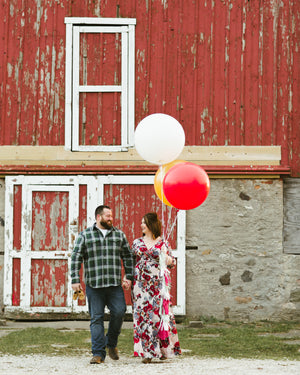 Couple standing in front of rustic red barn holding three jumbo balloons one in white, one in red, and one in gold.