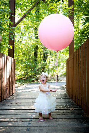 A little girl wearing a white dress holding a jumbo rose colored balloon.