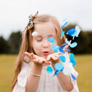 A little girl stands in a grass field blowing blue colored confetti from her hands with her eyes closed.