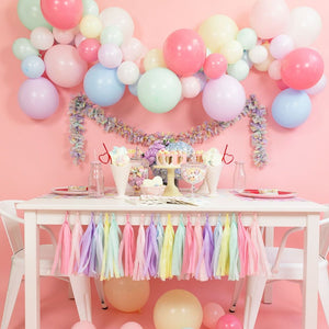 A cute pastel party decor set up with pastel colored balloon arch garland hanging from the wall. There is a pastel colored tissue tassel banner hanging from the white table with two white chairs. There are pastel colored balloons on the floor.