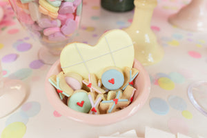 Pile of pastel colored hand cut tissue confetti paper on a pink table with bowl of pastel colored cookies on top.