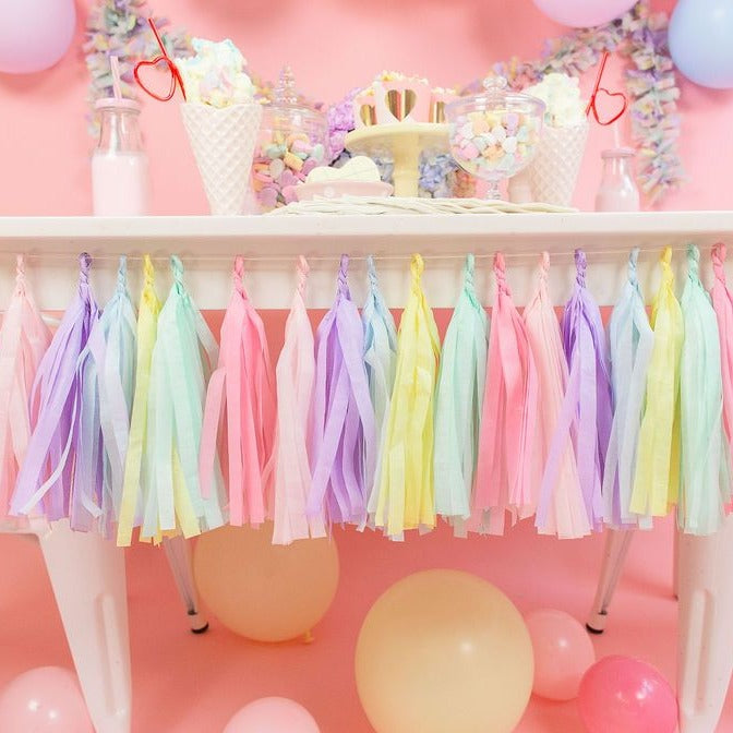 Pastel tissue garland with the colors pink, blush, lilac, light blue, light yellow, and mint hang from a white table with a pink backdrop. There are yellow and light pink balloons on the floor.