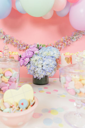 Pile of pastel colored hand cut tissue confetti paper on a pink table with a pastel garland on top.