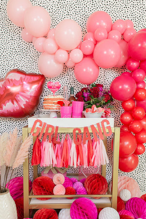 Valentines party decor with one jumbo 30 inch red and pink lip balloon, a balloon arch garland with different shades of pink and red, one BAE MINE banner, and a tissue tassel garland strung on a mini cart.