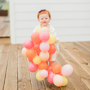 A little girl stands on a wooden deck outside holding up a 5 inch mini balloon garland made of goldenrod, coral, rose, and pink balloons.