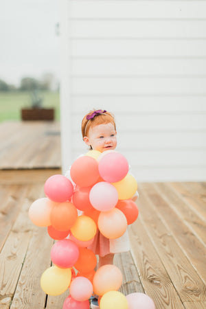 A little girl stands on a wooden deck outside holding up a 5 inch mini balloon garland made of goldenrod, coral, rose, and pink balloons.