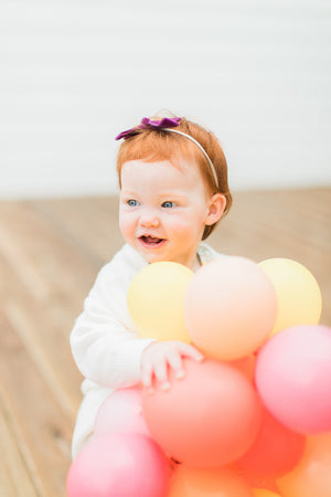 A little girl stands on a wooden deck outside holding up a 5 inch mini balloon garland made of goldenrod, coral, rose, and pink balloons.