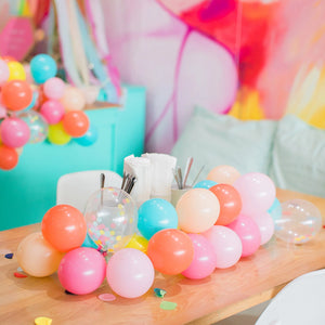 A bright mini balloon garland made up of only 5 inch balloons sits running along the top of a wooden table. The colors are coral, rose, blush, Caribbean blue, wildberry, yellow, and matching clear filled confetti balloons.