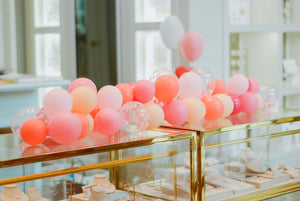 A mini balloon garland made up of rose, coral, pink, and blush sits top a gold metal framed jewelry display case.