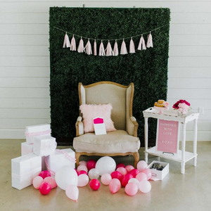Various inflated pink and white 5 inch balloons scattered on the ground in front a grass backdrop next to a tan colored chair, white side table, and white colored gift wrapped boxes.