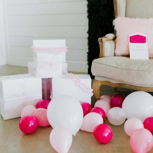 Various inflated pink and white 5 inch balloons scattered on the ground next to a tan colored chair and white gift wrapped boxes.