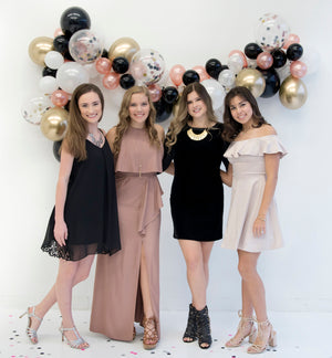 Four girls in dresses stand beside each other with arms around each other in front of the balloon garland smiling.