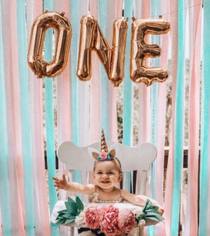 Little girl sitting in a high chair with rose gold mylar balloons spelling ONE hanging above her.