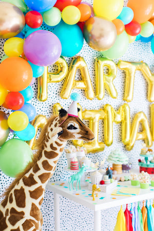 Party animal backdrop features giant gold letter balloons spelling PARTY ANIMAL, a colorful tissue garland hanging from a white table, and a rainbow colored balloon arch garland.