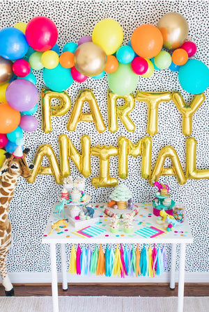 Party animal backdrop features giant gold letter balloons spelling PARTY ANIMAL, a colorful tissue garland hanging from a white table, and a rainbow colored balloon arch garland.
