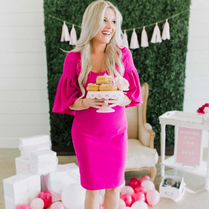 A woman wearing a hot pink dress holding a cake platter topped with donuts in front of a grass backdrop with various pink and white 5 inch balloons inflated on the ground around her.