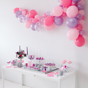 A pink and purple 10ft long balloon garland arch hangs above a white table that is full of pink and purple sweets and treats. There are cake pops, cookies, cotton candy, and purple colored drinks.