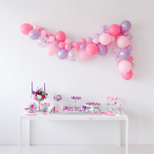 A frontal view of a pink and purple decorated party. There is a long balloon garland hanging on a white wall with a white table below it full of pink and purple sweets and treats.