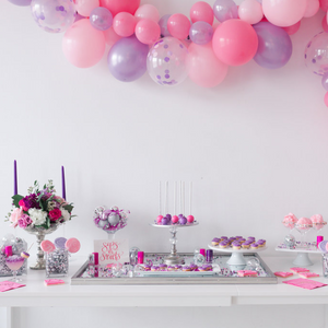 Close up of a white table full of pink and purple sweets and treats. Above the table is a pink and purple balloon garland.