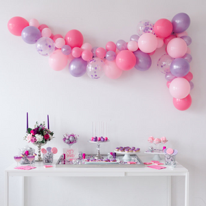 A picture perfect pink and purple party theme. There is a pink and purple balloon garland hanging above a white table full of pink and purple decorated sweets and treats. 