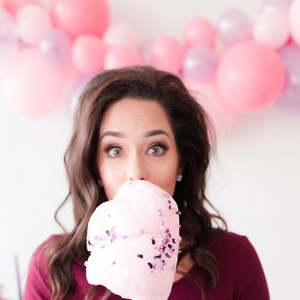 A woman is looking at the camera holding pink cotton candy in front of her face. Behind her is a pink and purple balloon garland.