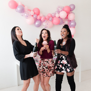 Three woman are popping champagne bottles in excitement. Behind them is a pink and purple balloon garland.