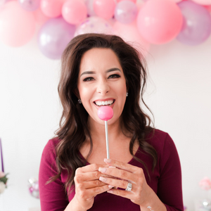 A woman is looking at the camera holding a pink cake pop in front of her face and smiling. Behind her is a pink and balloon garland hanging from a white wall.