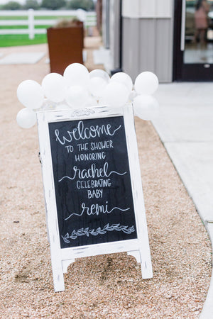 A black chalk sign sits outside welcoming guest to a baby shower and a white mini balloon garland sits on top of it.