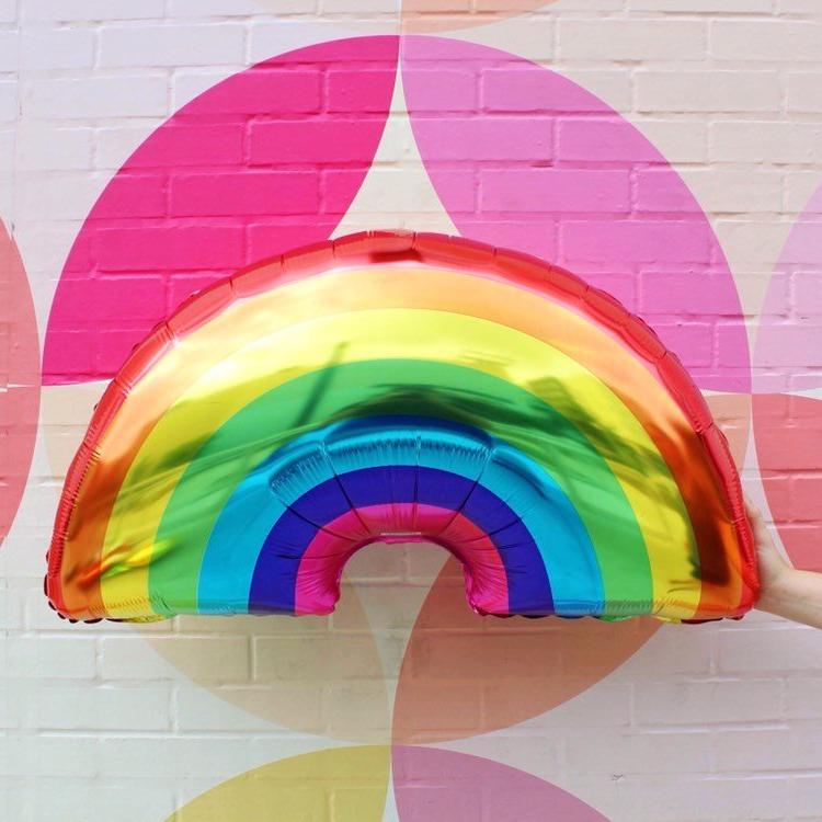 Womans hand holding a jumbo Rainbow Party Balloon in front of an abstract painted wall.