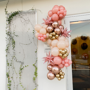 A pink and chrome balloon garland hangs next to the front door of a boutique. The balloon garland includes rose gold starburst balloons.