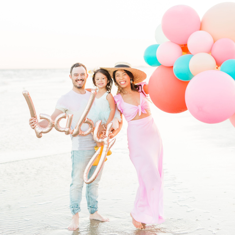 Couple holding their toddler and a rose gold script BABY balloon at the beach.