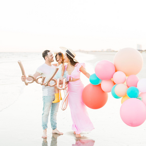 Couple at the beach holding their baby girl and a rose gold script BABY balloon. The woman is also holding an assortment of balloons on her left hand.