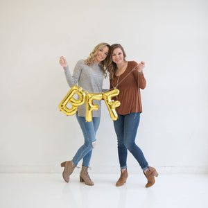 Two girls stand hugging each other and facing the camera while holding gold letters BFF on a string of twine.