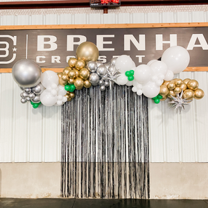 A long balloon garland hangs as a backdrop at an event. The balloons are in the colors chrome silver, chrome gold, white, and green. There are also silver starburst balloons in the balloon garland.