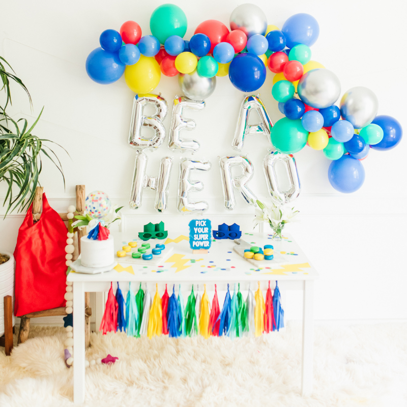 A colorful balloon garlands hangs on top of jumbo silver letters that spell BE A HERO. Below sits a white table with superhero themed birthday party sweets and decorations.