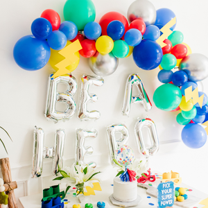 A close up view of a colorful balloon garland with paper lighting bolts and silver jumbo balloons that say BE A HERO. Below is a white table with a white birthday cake surrounded by superhero themed sweets and decorations.