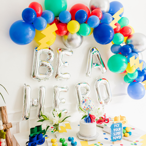 A balloon garland with the colors blue, yellow, green and red is hung on a white wall. Below the garland are silver jumbo balloon letters that read BE A HERO. Below the balloons is a white table with matching superhero themed birthday sweets and decorations.