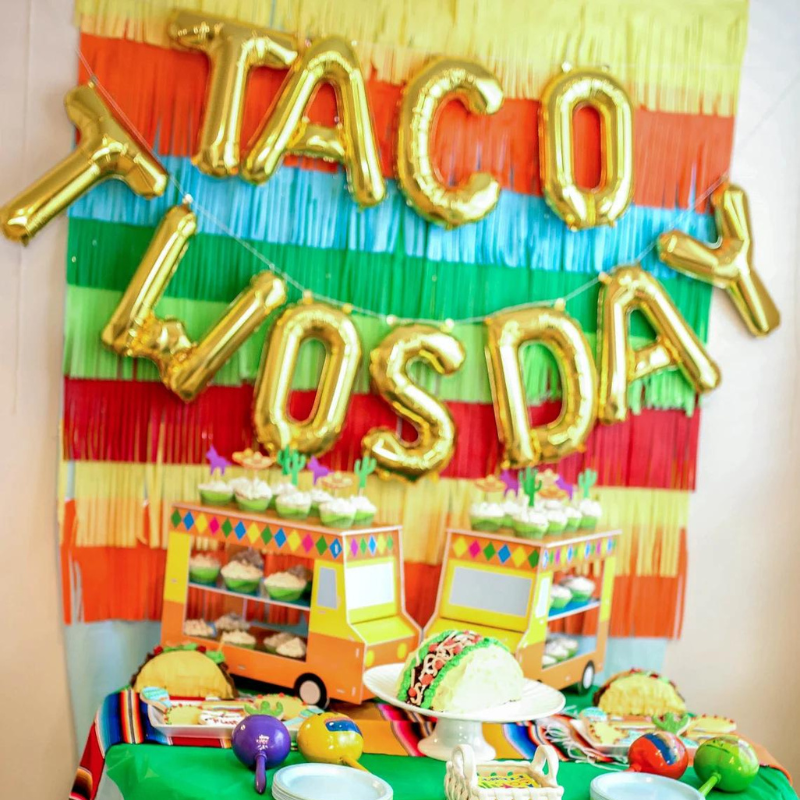 Two year olds birthday party with a fiesta theme. There are gold balloon letters hanging on a backdrop spelling TACO TWOSDAY. On the table there is a taco cake, taco pinatas, and more fiesta themed decorations!