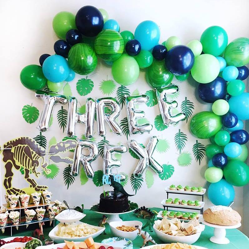 A three year olds dinosaur theme birthday party. There are silver jumbo letter balloons spelling THREE rex and a balloon arch above it in the colors green and blue.