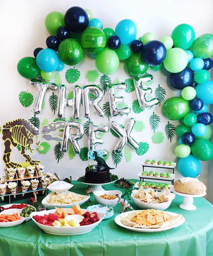 A three year olds dinosaur theme birthday party. There are silver jumbo letter balloons spelling THREE rex and a balloon arch above it in the colors green and blue.