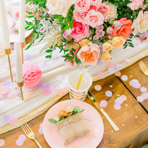 An arial shot shows soft pink, lilac, and blush one inch circular tissue confetti spread all over a wooden table with a pretty bouquet of flowers sitting in the middle.