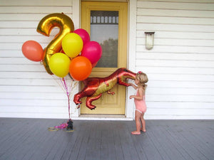 A little girl on the front porch of a house with a bouquet of balloons in yellow, coral, orange, wildberry, a number 2 in gold, and dinosaur balloon.