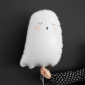 A womans hand wearing a black and white polka dot long sleeve is holding a 26 inch tall white ghost balloon in front of a dark gray wall.