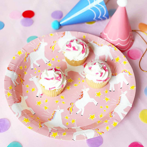Pink plate with white unicorn design and gold stars on a pink birthday table. There are three white frosting cupcakes on the plate and two party hats next to it.