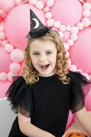 A little girl is looking at the camera wearing a black witches costume with a black witch party hat. Behind her is a wall of pink balloons.