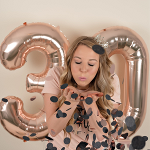 A woman blows black confetti towards the camera with her eyes closed. Behind her are two rose gold jumbo number balloons making a 30.