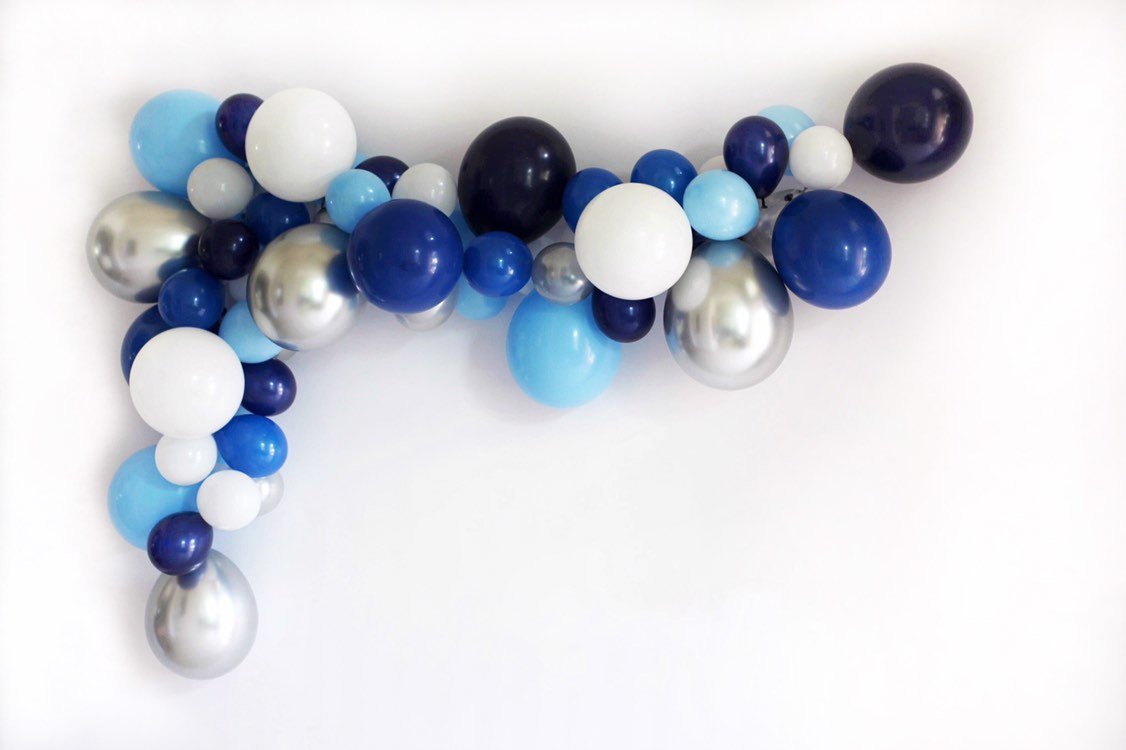 A wintery themed balloon garland is draped on a white wall in a mix of white, silver, chrome silver, light blue, blue, and navy 5 inch and 11 inch balloons.